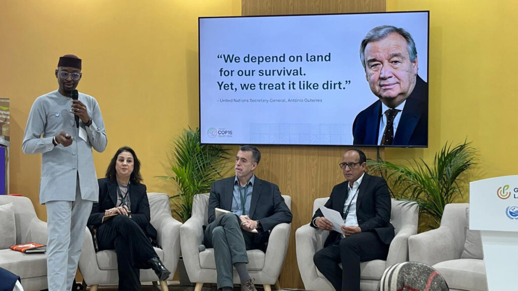 A presenter standing in front of an audience, with other panel members at the background, during an event at the Restoration Pavilion, on 4 December 2024, in Riyadh, Saudi Arabia